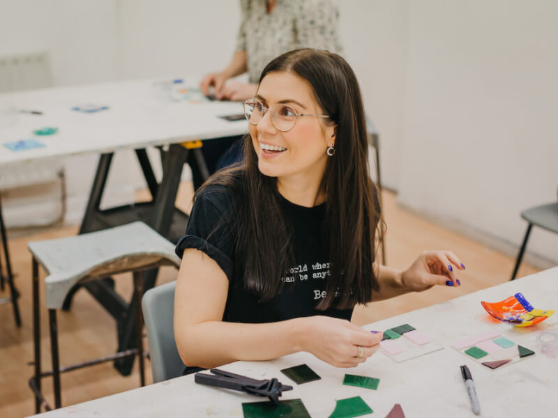 Woman in glasses smiling as they create mosaic art with pink and green glass.